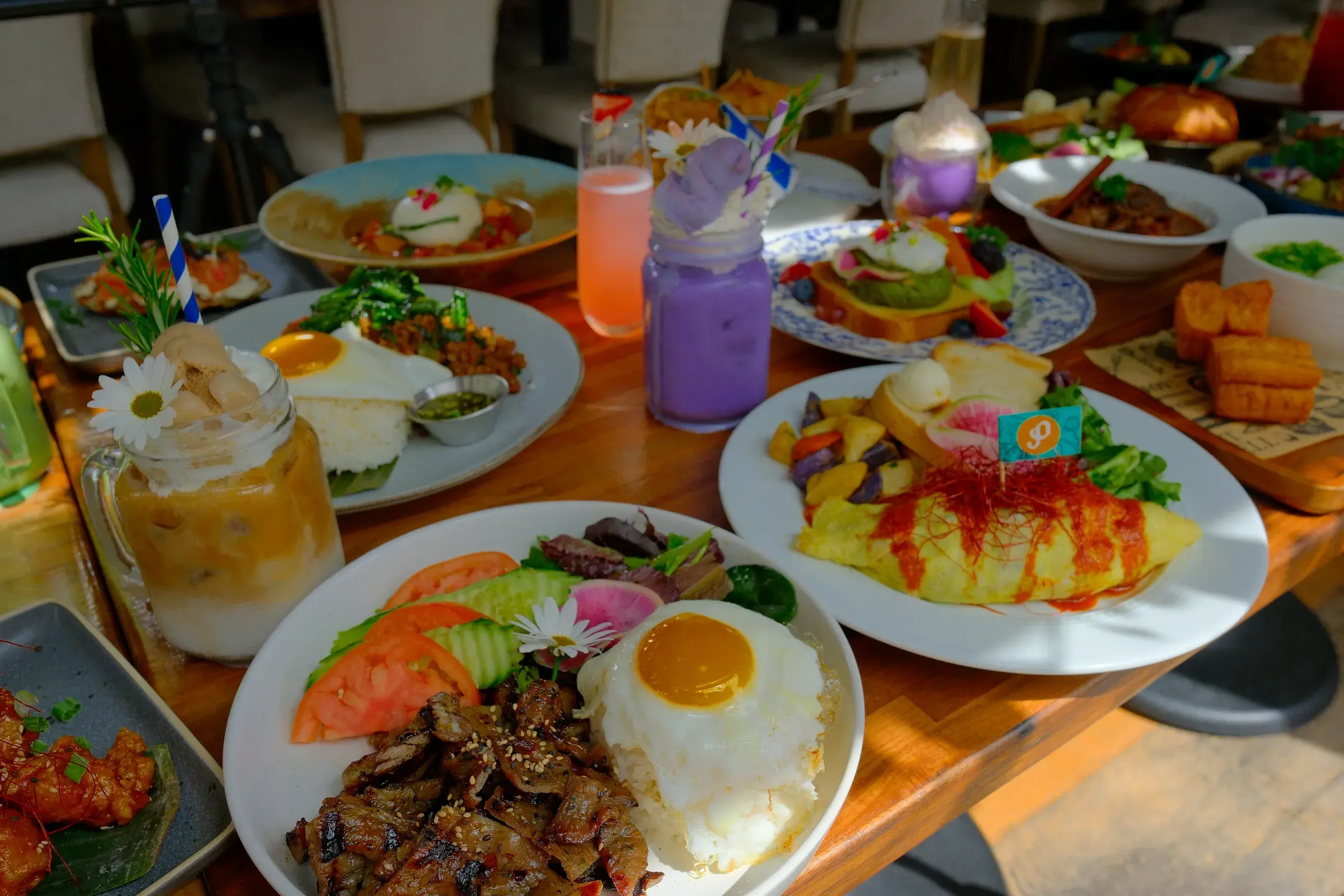 A wooden table of brunch plates: a fried egg over grilled meat and rice, an omelette, salmon toast and iced drinks
